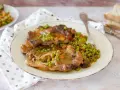 Un piatto di ossobuco con piselli e carote, servito su un piatto bianco con una forchetta. Sullo sfondo, fette di pane.