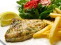 A cooked fish fillet with french fries, salad, and lemon slices on a white plate.