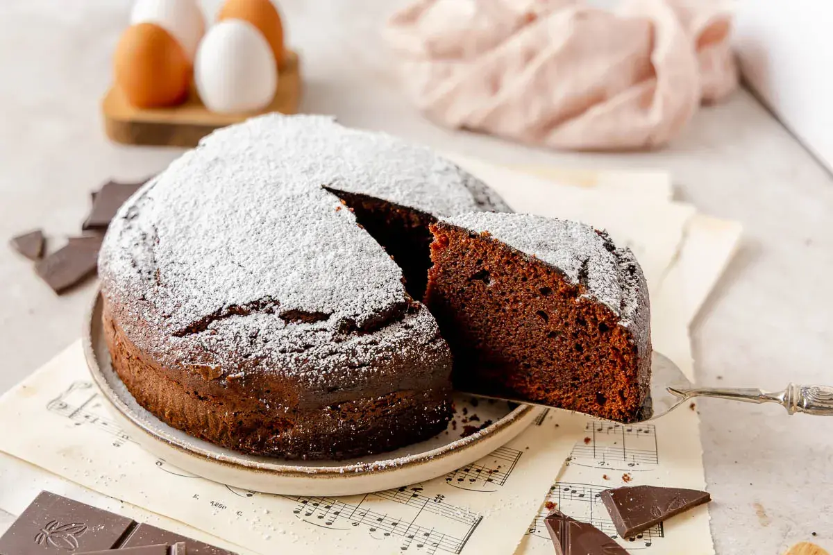 Una fetta di torta al cioccolato, soffice e spolverata di zucchero a velo, viene sollevata da una paletta. Pezzi di cioccolato e uova sullo sfondo.