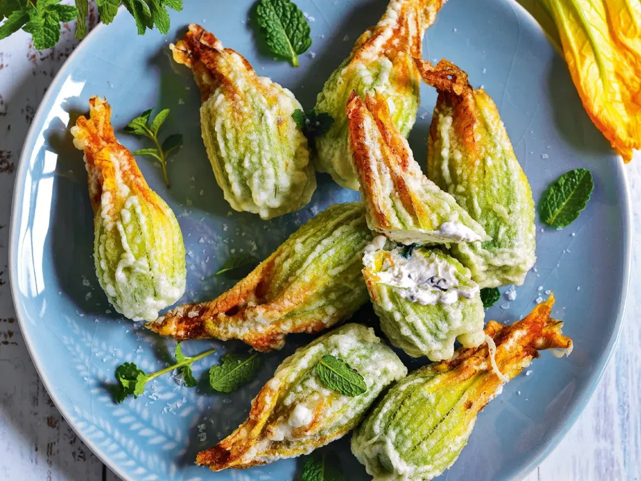Fiori di zucca fritti, ripieni di formaggio cremoso e guarniti con foglie di menta fresca, su un piatto blu.