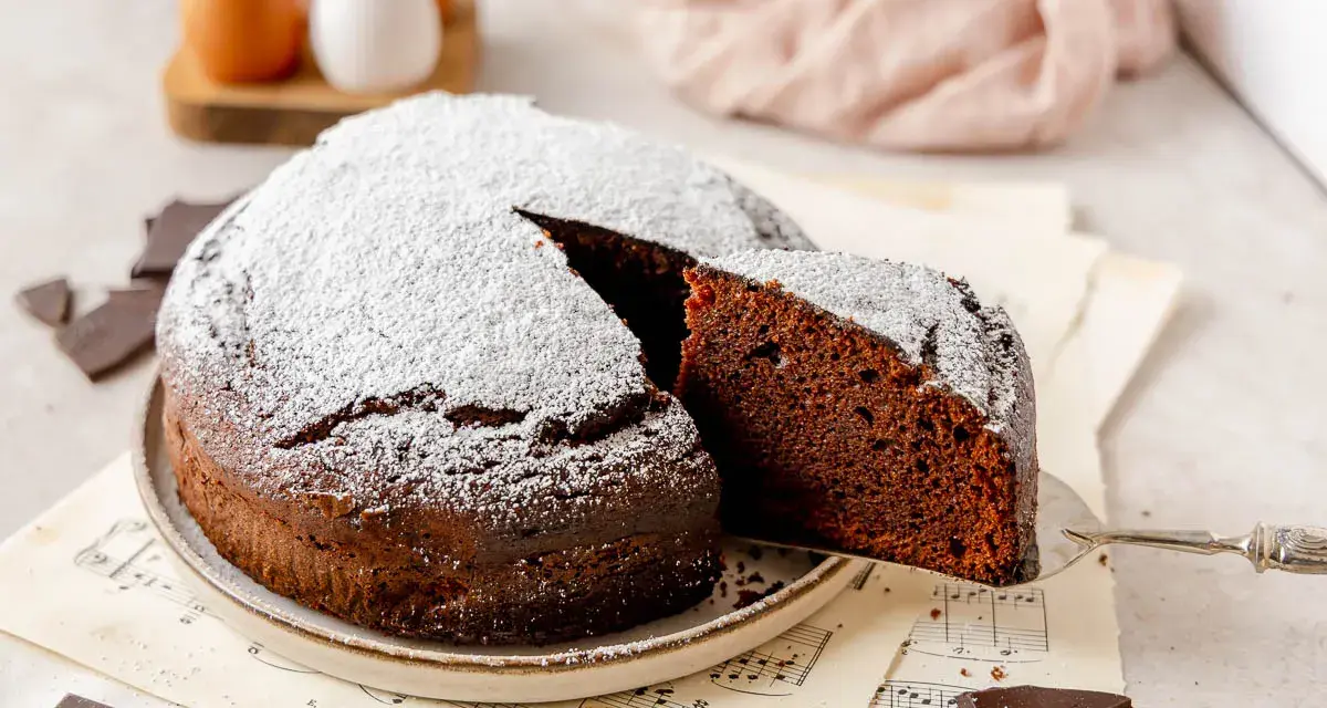 Una fetta di torta al cioccolato, soffice e spolverata di zucchero a velo, viene sollevata da una paletta. Pezzi di cioccolato e uova sullo sfondo.