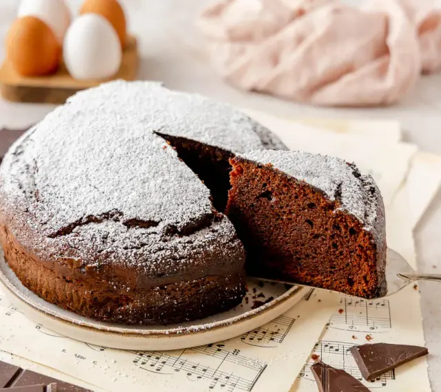 Una fetta di torta al cioccolato, soffice e spolverata di zucchero a velo, viene sollevata da una paletta. Pezzi di cioccolato e uova sullo sfondo.