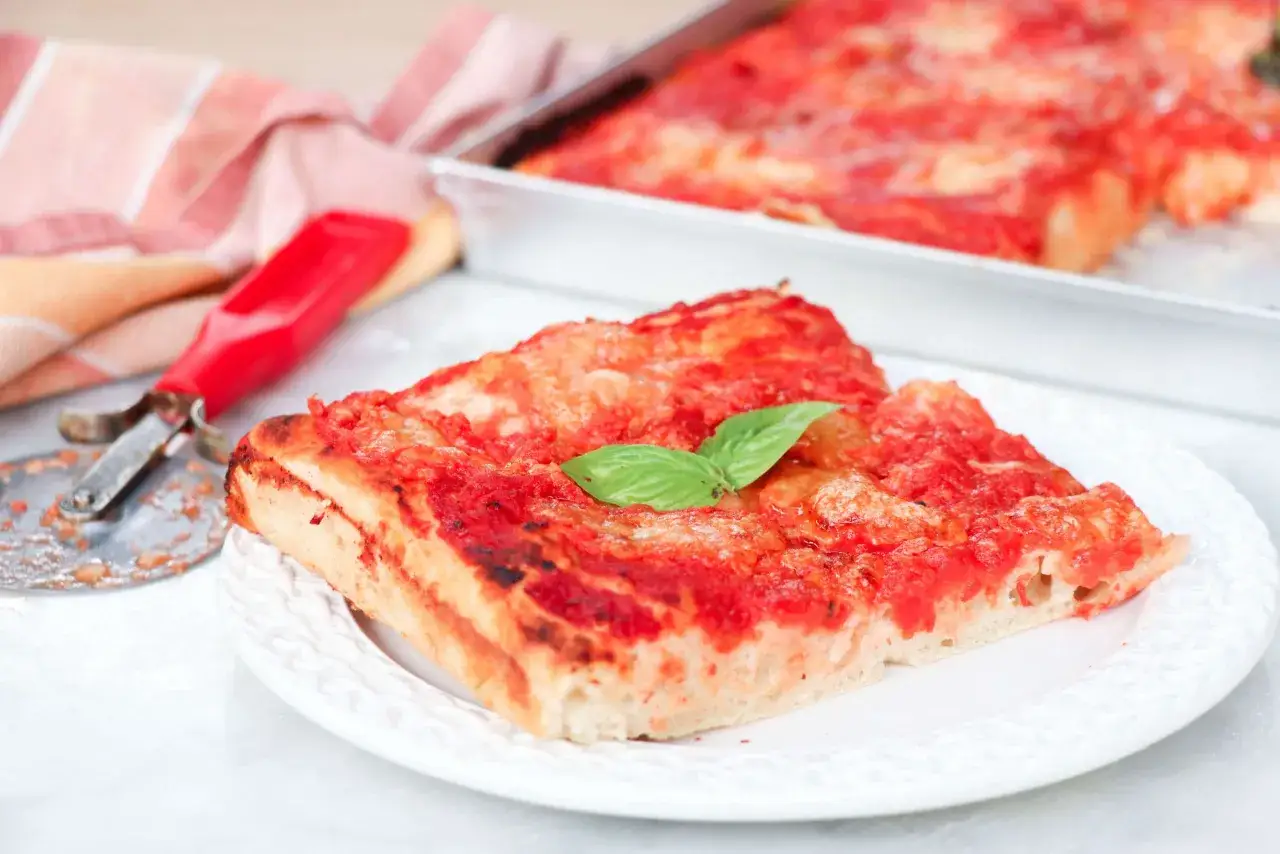 Una fetta di pizza fatta in casa, con pomodoro e basilico fresco, su un piatto bianco. Accanto, un tagliapizza rosso e un tovagliolo a righe.