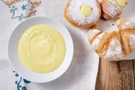 Una ciotola di soffice crema pasticcera senza uova accanto a cornetti e bomboloni spolverati di zucchero a velo.