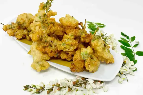 Fiori di acacia fritti, dorati e croccanti, serviti su un piatto bianco con rametti di fiori e foglie fresche.