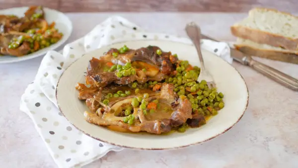 Un piatto di ossobuco con piselli e carote, servito su un piatto bianco con una forchetta. Sullo sfondo, fette di pane.