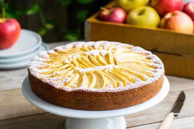 Torta di mele velocissima in 10 minuti, decorata con fettine di mela e zucchero a velo. Sullo sfondo, una cassetta di mele rosse e gialle.