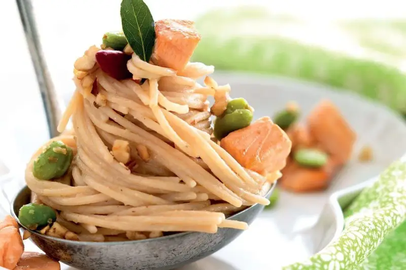Spaghetti di riso con salmone, fave e noci, guarniti con una foglia verde. Un piatto invitante e colorato.