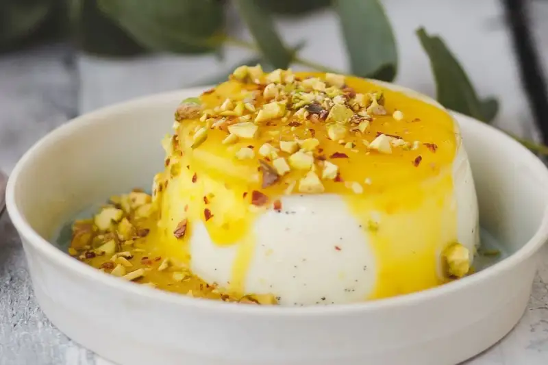 Budino fatto in casa cremoso, con salsa gialla e granella di pistacchi, servito in una ciotola bianca.
