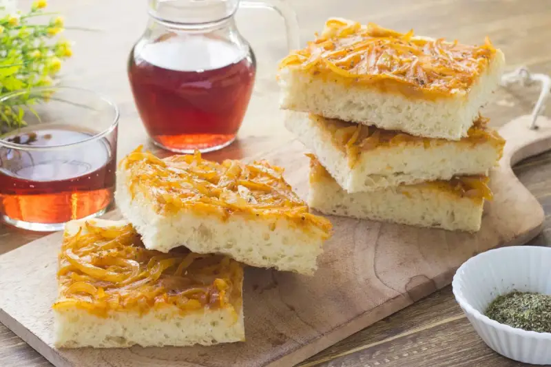 Focaccia con cipolle dorate e soffice, servita su un tagliere di legno con un bicchiere di vino rosato.